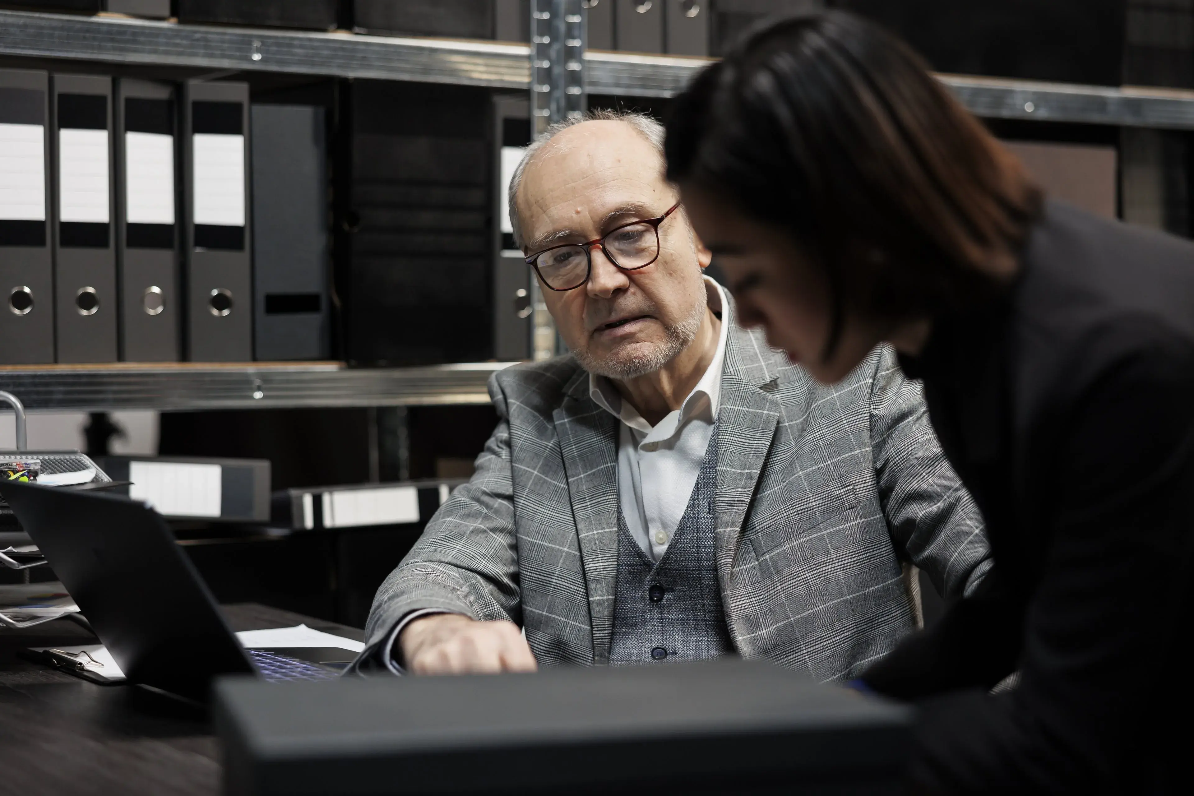 Two professionals reviewing records on a laptop in an office lined with files